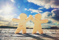 Wooden little men holding hands on sky and sun background. Symbol of friendship, love and teamwork