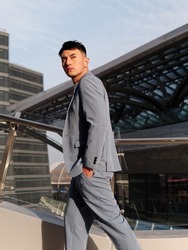 Portrait of handsome Chinese young man in light blue suit and white undershirt walking and looking away with modern city buildings background in sunny day, side view of confident businessman.