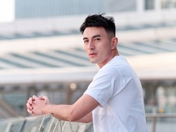 Portrait of handsome Chinese young man in white T-shirt standing and looking at camera with modern city buildings background in sunny day, side view of confident young man.