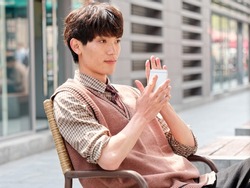 Portrait of handsome Chinese young man with curly black hair in plaid shirt and wool vest sitting outdoor cafe with his mobile phone in hand in sunny day, male fashion, cool Asian young man lifestyle