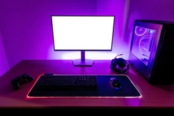 Desk with gaming setup. Display with isolated screen for mockup. Gaming PC, headset, keyboard, mouse and joypad on desk. Purple led light on wall