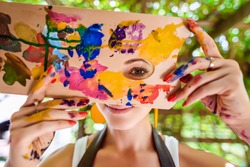 Playful portrait of a young gorgeous female artist painter covered in paint, looking and smiling at camera through her painter's palette. Creativity and individuality concept.