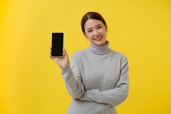 Portrait of female Asian tourist, holding blank template mobile phone for presenting mobile application, grey shirt, isolated on yellow background