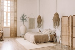 Spacious, bright bedroom with white walls and a large bed in warm boho tones. Straw chandeliers, a large decorative vase, wooden shutters on the windows.