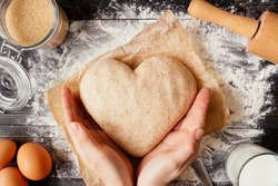 female hands holding dough in heart shape top view. Baking ingredients on the dark wooden table