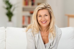 Lovely middle-aged blond woman with a beaming smile sitting on a sofa at home looking at the camera