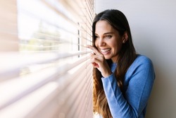 Smiling young woman peeking looking through venetian blinds on window at home