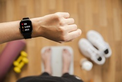Looking at fitness tracker displaying heart rate, steps count and burnt calories while standing on scales, first person view. Smart watch on female hand, point of view, concept of staying fit at home