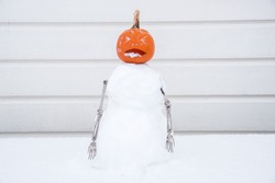 Snowman with Jack-o-lantern pumpkin head  and skeleton arms making scary faces in the snow against white wooden wall background.