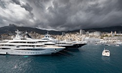 A lot of huge yachts are in port of Monaco at storm weather, mountain is on background, glossy board of the motor boat, megayachts are moored in marina, sun reflection on glossy board