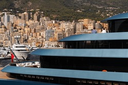 Landmarks of Monaco through decks of huge yacht of blue color in port Hercule at sunny day, glossy board of the motor boat, sun reflection on glossy board, mountains are on background
