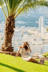 a bright beautiful girl in a light dress and hat sits on the grass under a palm tree in Monaco in sunny weather in summer, streets of old town of Monaco