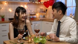 smiling asian young lovers wearing suit and dress eating romantic dinner together with fun chat in celebration of valentine’s day in a cozy home interior
