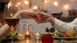 closeup with cropped shot of lovers holding hands over romantic valentine’s day dinner table with wine and red rose. the man gently touches the woman’s fingers.