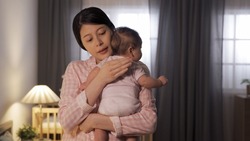 waist up asian mom is embracing and patting on her baby’s back trying to put her to sleep at nighttime in illuminated home interior.