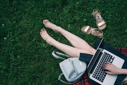 Top view of woman sitting in park on the green grass with laptop, notebook and smartphone, hands on keyboard. Computer screen mockup. Student studying outdoors. Copy space for text
