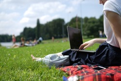 Young woman's legs on the green grass with open laptop. Girl's hands on keyboard. Distance learning concept