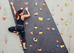 Female fitness professional climber training at bouldering gym. Muscular woman with athletic body dressed in black, climbing on artificial colourful rock wall. Active lifestyle and bouldering concept.