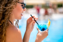 close up face profile portrait of young beautiful caucasian woman with long brown hair wearing sunglasses drinking blue cocktail though a straw over outdoor poolside area blurred bckground.