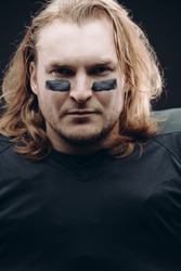 Portrait of determined caucasian blonde American football sportsman in black uniform with painted black stripes under his eyes on his face posing against black background