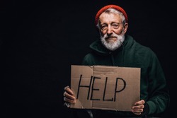Sad exhausted old-aged bum in red hat and street wear holding a cardboard sign, seeking help, job and food posing at studio over black wall.