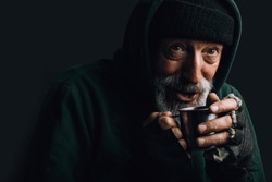 Old homeless man with grey beard covering up in green decrepit wear holding a mug of hot tea to warm himself in a cold night
