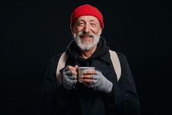 Bearded beggar male smiling, happy even living in street. Cheerful homeless man with mug of hot tea isolated over black background