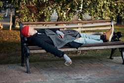 Homeless man sleeping on bench in the park during day. Mature grey bearded man in street clothes. Side view