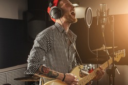 young caucasian gorgeous musician play electric guitar and sing on microphone in recording studio. rockstar perform music. man preparing, practicing before concert.