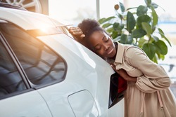 attractive afro woman dreams about new car, young female came to see automobiles, make purchase. woman is leaned on white luxurious car, smiling