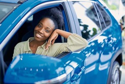 satisfied african woman behind the wheel of blue automobile represented in cars showroom, she looks at camera and smile