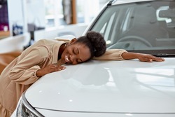 smiling black lady hugs her new auto in cars showroom. young woman fulfill her dream, lies on car
