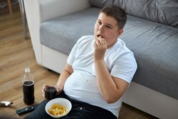 overweight fat boy eat junk food while watching tv alone at home, sit on the floor with french fries, carbonated drink, chips. enjoy unhealthy lifestyle