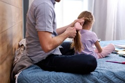 Father braiding daughters hair carefully, taking care of beloved child girl, family in casual wear sit on bed at home, preparing for school in morning