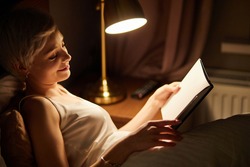 Beautiful female in pajamas lying on bed reading book, alone at night. Caucasian short haired lady in bedroom, charming cute woman in room lighted by lamp. side view