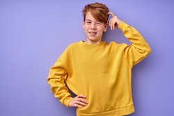 Portrait of pensive teen boy isolated on purple background. Thoughtful teenager holding hand on head, looking at camera. Beautiful caucasian funny child in shirt is thinking, in contemplation