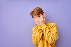 Portrait of scared caucasian teen boy, isolated on purple studio background. Frightened child looking at camera through fingers, closing eyes, in shock or amazement. human emotions, people concept