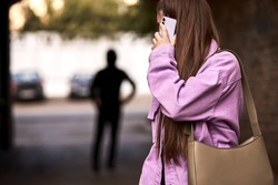 Criminal stalking woman, commiting crime while victim was walking alone, talking on phone in dark street. Caucasian young woman is looking back, afraid of male stranger person in the background