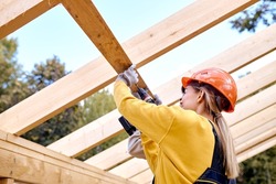 Two Positive Contractors Working With Drill Driver. Carpenter Working on Wooden House Skeleton Frame Roof Section. Construction Industry Theme. Woman In Hardhat Is Laughing During Work