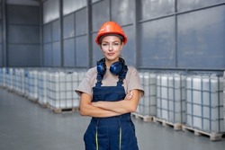 industrial woman engineer in hardhat standing in factory with arms folded, posing, wearing uniform, at work place, caucasian lady is smiling. production, engineering, industry concept