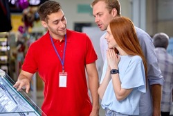 Fiendly Consultant Help Customers Couple In Store, Make Choice, Buy Smartphone, Talking, Having Discussion. Male Consultant In Red Uniform Working With Clients Talk About Models Options