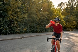 Professional fit cyclist in black helmet, protective glasses and active wear dynamically riding bicycle drinking water for better speed. Male preparing for competitions and races on fresh air