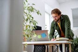 Bearded young confident guy stand behind office desk with laptop thinking working alone. handsome male in casual wear engaged in office work, planning strategy, deadlines. at workplace