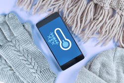 Concept for cold temperatures with snow and minus degrees with mobile phone showing weather forecast surrounded by warm winter clothes like scarf, hat and gloves