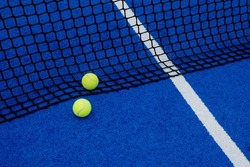 Two balls near the net of a blue artificial grass paddle tennis court.