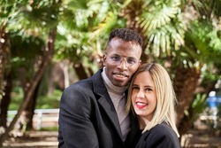 Black man and blond woman looking at camera while standing against palms during romantic date on sunny day in exotic park