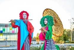 Full length of creative talented performers with face art in colorful costumes and wigs standing on stilts in park with Asian umbrella and sword during show, on sunny day