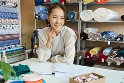 Portrait of a smiling Asian woman seamstress in her workshop against the background of shelving with fabrics