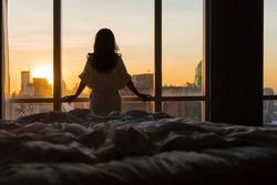 Beautiful asian woman is waking up in the morning, Sun shines on her from the big window. Happy young girl greets new day with warm sunlight flare and city scenery in the window. Co