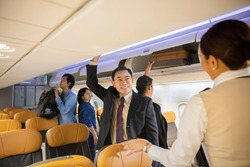 Group of passengers putting luggage into overhead locker on airplane. Traveler placing carry on bag in overhead compartment in aircraft while Flight attendant woman service helping passenger.  